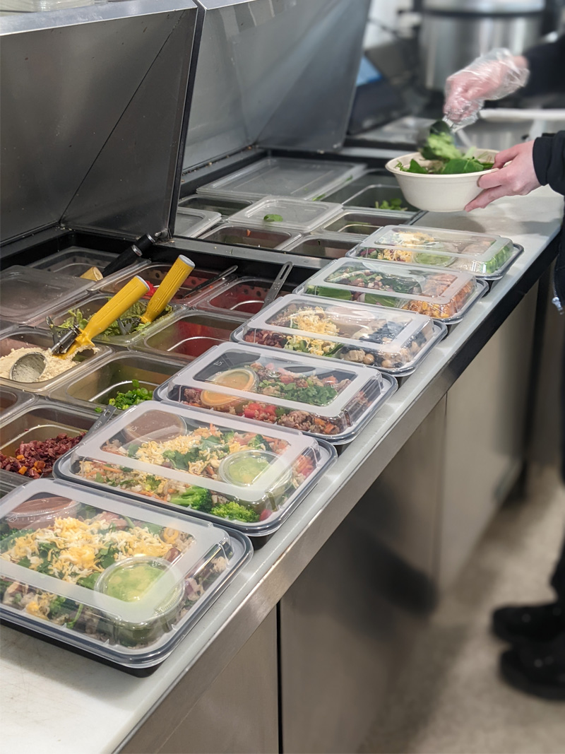 A row of fresh healthy meal containers being prepared with protein, grains, and vegetables for the grab-and-go section at All Good Eats & Drinks.